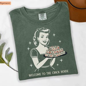 Op de afbeelding: Blauwspar-kleurig T-shirt met een retro illustratie van een vrouw die een dienblad met Mahjong-tegels vasthoudt. De tekst "WELCOME TO THE CRACK HOUSE" staat onderaan. Het shirt is van het merk Comfort Colors.