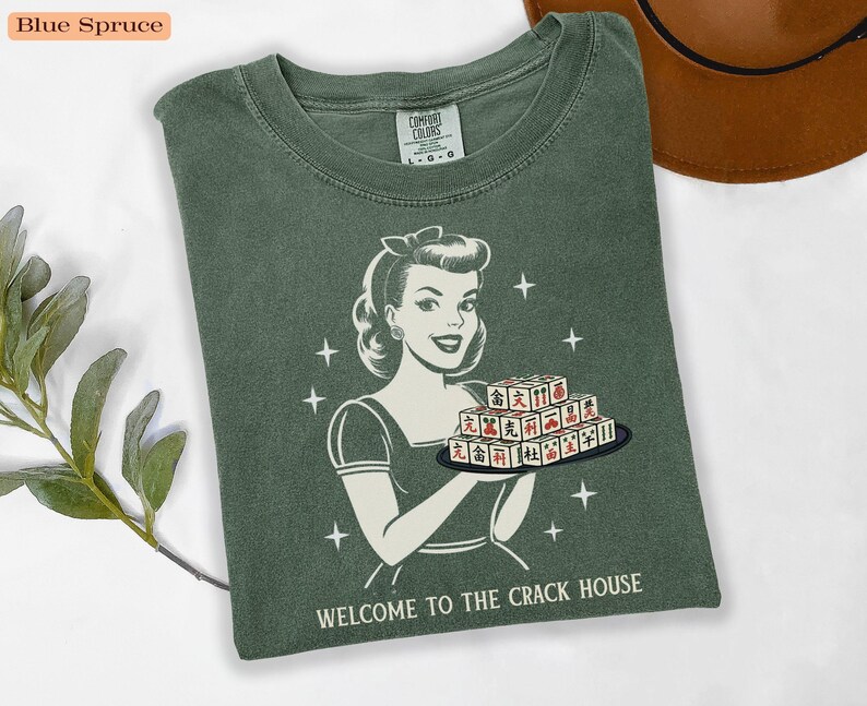 Op de afbeelding: Blauwspar-kleurig T-shirt met een retro illustratie van een vrouw die een dienblad met Mahjong-tegels vasthoudt. De tekst "WELCOME TO THE CRACK HOUSE" staat onderaan. Het shirt is van het merk Comfort Colors.