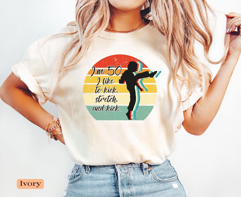Puede incluir: Una camiseta de color marfil con un gr&aacute;fico circular de rayas horizontales en rojo, amarillo y verde azulado. El dise&ntilde;o incluye la silueta negra de una persona dando una patada alta y el texto: I'm 50 I like to kick. stretch. and kick.