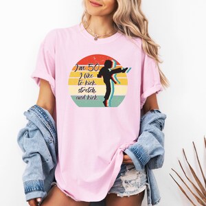 Puede incluir: Una camiseta rosa claro con un gr&aacute;fico circular que muestra un atardecer retro en amarillo, naranja y turquesa. El texto dice "I'm 50 I like to kick, stretch, and kick." Una silueta negra de una persona dando una patada alta.