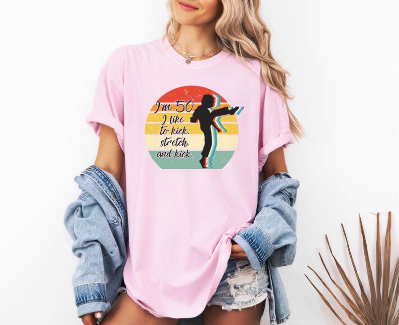 Puede incluir: Una camiseta rosa claro con un gr&aacute;fico circular que muestra un atardecer retro en amarillo, naranja y turquesa. El texto dice "I'm 50 I like to kick, stretch, and kick." Una silueta negra de una persona dando una patada alta.