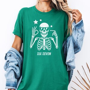 以下が含まれることがあります： サングラスをかけた白い骸骨のグラフィックが特徴の緑色のTシャツ。骸骨の下には「SIX SEVEN」の文字。骸骨の頭の上には2つの星があります。