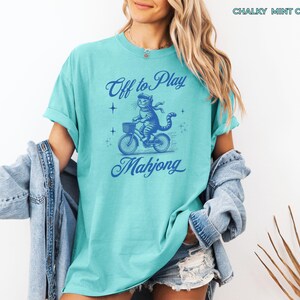 May include: A light teal t-shirt with a blue graphic of a cat riding a bicycle with a basket. The text "Off to Play Mahjong" is above and below the cat illustration. The shirt is a chalky mint color.