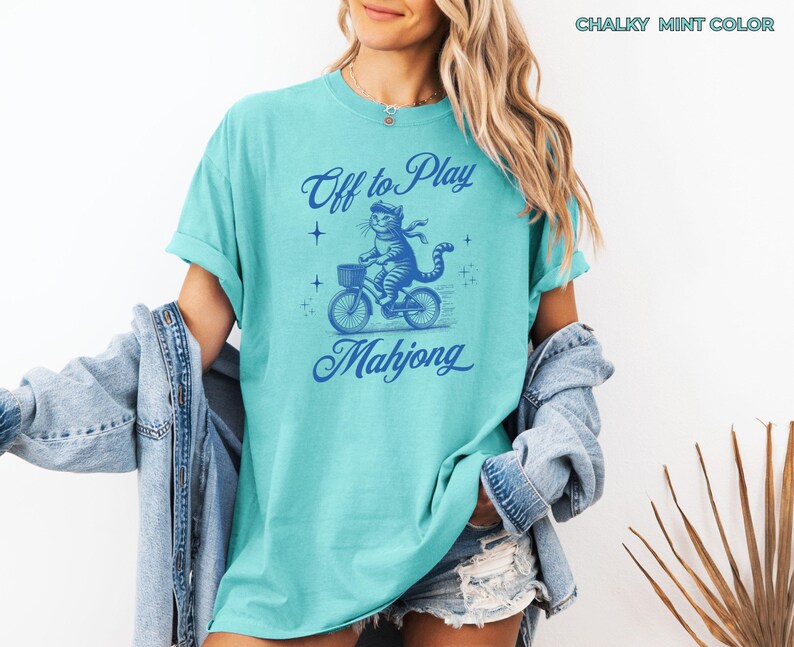 May include: A light teal t-shirt with a blue graphic of a cat riding a bicycle with a basket. The text "Off to Play Mahjong" is above and below the cat illustration. The shirt is a chalky mint color.
