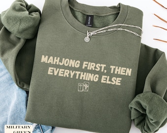 May include: A military green crewneck sweatshirt featuring the beige text "MAHJONG FIRST, THEN EVERYTHING ELSE" printed on the chest, accompanied by two small line-art illustrations of mahjong tiles positioned below the text.