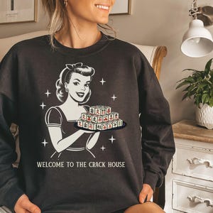 Op de afbeelding: Donkergrijze sweatshirt met een retro illustratie van een vrouw die Mahjong-tegels vasthoudt. De witte afbeelding bevat de tekst "WELCOME TO THE CRACK HOUSE". De sweatshirt heeft een ronde hals en is een casual kledingstuk.
