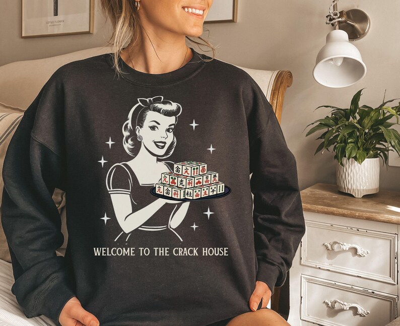 Op de afbeelding: Donkergrijze sweatshirt met een retro illustratie van een vrouw die Mahjong-tegels vasthoudt. De witte afbeelding bevat de tekst "WELCOME TO THE CRACK HOUSE". De sweatshirt heeft een ronde hals en is een casual kledingstuk.