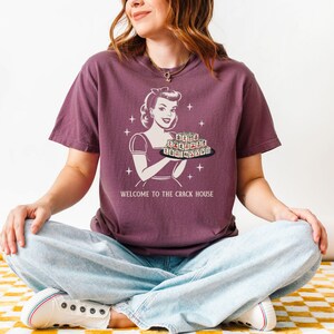 Op de afbeelding: Een bordeauxrood T-shirt met een retro-illustratie van een vrouw die een dienblad met letters vasthoudt. De tekst "WELCOME TO THE CRACK HOUSE" staat eronder. De persoon draagt een lichtblauwe jeans en witte sneakers.