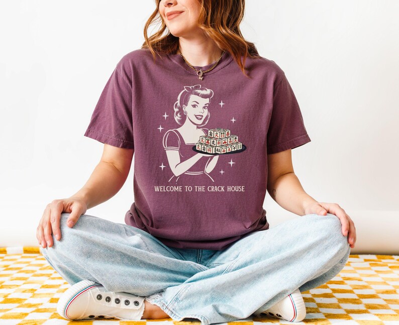 Op de afbeelding: Een bordeauxrood T-shirt met een retro-illustratie van een vrouw die een dienblad met letters vasthoudt. De tekst "WELCOME TO THE CRACK HOUSE" staat eronder. De persoon draagt een lichtblauwe jeans en witte sneakers.