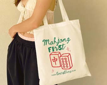 May include: A white canvas tote bag featuring a green and red graphic print. The text reads Mahjong FIRST then Everything ELSE, accompanied by illustrations of two mahjong tiles with red symbols and small star accents.