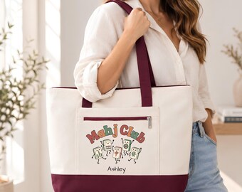 May include: A cream-colored canvas tote bag with a deep maroon bottom and matching shoulder straps. The front features a zippered pocket with the text "Mahj Club" above a graphic of four dancing mahjong tiles and the name "Ashley" below.