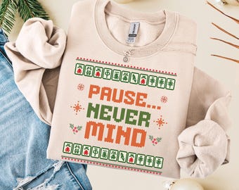 May include: A beige crewneck sweatshirt featuring a graphic print with the text PAUSE... NEVER MIND in orange and green. The design includes rows of mahjong tiles, snowflakes, and holly berries in red and green, creating a festive pattern.