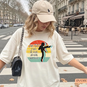 Puede incluir: Una camiseta color marfil con un gr&aacute;fico de una persona dando una patada alta dentro de un dise&ntilde;o circular de atardecer con el texto "I'm 50, I like to kick, stretch and kick." Se combina con una gorra de b&eacute;isbol beige y un bolso de hombro negro.
