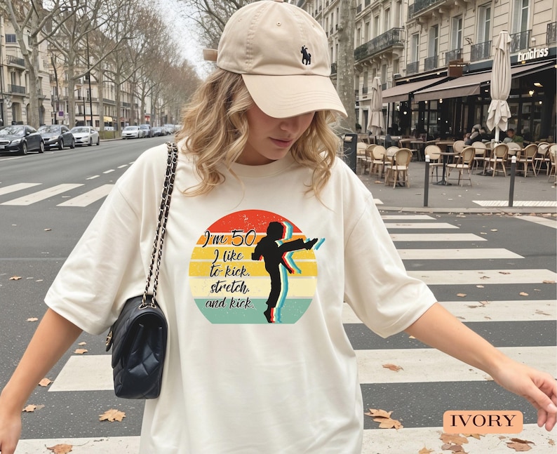 Puede incluir: Una camiseta color marfil con un gr&aacute;fico de una persona dando una patada alta dentro de un dise&ntilde;o circular de atardecer con el texto "I'm 50, I like to kick, stretch and kick." Se combina con una gorra de b&eacute;isbol beige y un bolso de hombro negro.