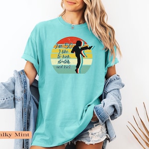Puede incluir: Una camiseta de manga corta color menta tiza con un gr&aacute;fico de una persona realizando una patada alta dentro de un c&iacute;rculo de estilo atardecer retro. El texto dice, I'm 50, I like to kick, stretch and kick. El dise&ntilde;o utiliza colores negro, rojo, amarillo y azul.