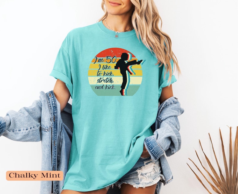 Puede incluir: Una camiseta de manga corta color menta tiza con un gr&aacute;fico de una persona realizando una patada alta dentro de un c&iacute;rculo de estilo atardecer retro. El texto dice, I'm 50, I like to kick, stretch and kick. El dise&ntilde;o utiliza colores negro, rojo, amarillo y azul.