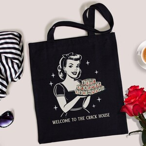 Op de afbeelding: Zwarte draagtas met een retro illustratie van een vrouw die een dienblad met tegels vasthoudt, met de tekst "WELCOME TO THE CRACK HOUSE". De tas heeft zwarte handvatten. Een zwart-witte sjaal, zonnebril, kopje koffie en rode rozen zijn ook zichtbaar.
