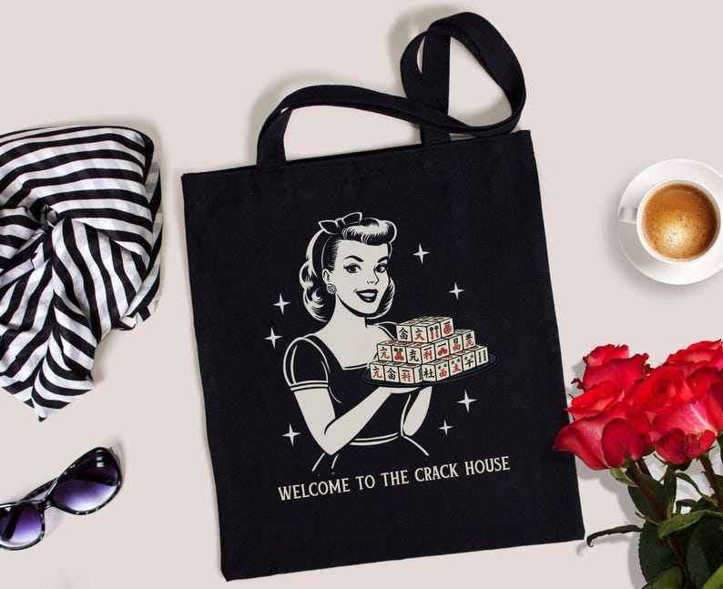 Op de afbeelding: Zwarte draagtas met een retro illustratie van een vrouw die een dienblad met tegels vasthoudt, met de tekst "WELCOME TO THE CRACK HOUSE". De tas heeft zwarte handvatten. Een zwart-witte sjaal, zonnebril, kopje koffie en rode rozen zijn ook zichtbaar.