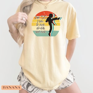 Puede incluir: Una camiseta de manga corta de color amarillo p&aacute;lido con un gr&aacute;fico circular de un atardecer retro en rojo, amarillo y turquesa. El gr&aacute;fico incluye la silueta negra de una persona dando una patada y el texto: I'm 50 I like to kick, stretch, and kick.