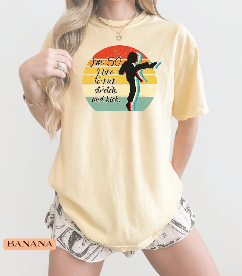 Puede incluir: Una camiseta de manga corta de color amarillo p&aacute;lido con un gr&aacute;fico circular de un atardecer retro en rojo, amarillo y turquesa. El gr&aacute;fico incluye la silueta negra de una persona dando una patada y el texto: I'm 50 I like to kick, stretch, and kick.