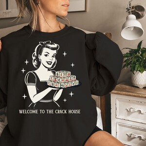 Op de afbeelding: Zwart sweatshirt met een retro illustratie van een vrouw die een dienblad met Mahjong-tegels vasthoudt. De afbeelding is in zwart-wit met rode accenten op de tegels. De tekst "WELCOME TO THE CRACK HOUSE" staat eronder.