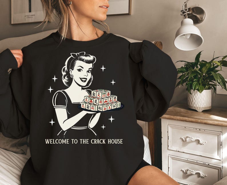 Op de afbeelding: Zwart sweatshirt met een retro illustratie van een vrouw die een dienblad met Mahjong-tegels vasthoudt. De afbeelding is in zwart-wit met rode accenten op de tegels. De tekst "WELCOME TO THE CRACK HOUSE" staat eronder.