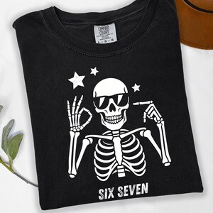 以下が含まれることがあります： サングラスをかけた白い骸骨のグラフィックが特徴の黒いTシャツ。「SIX SEVEN」の文字が骸骨の下にプリントされています。Tシャツはクルーネックで半袖です。