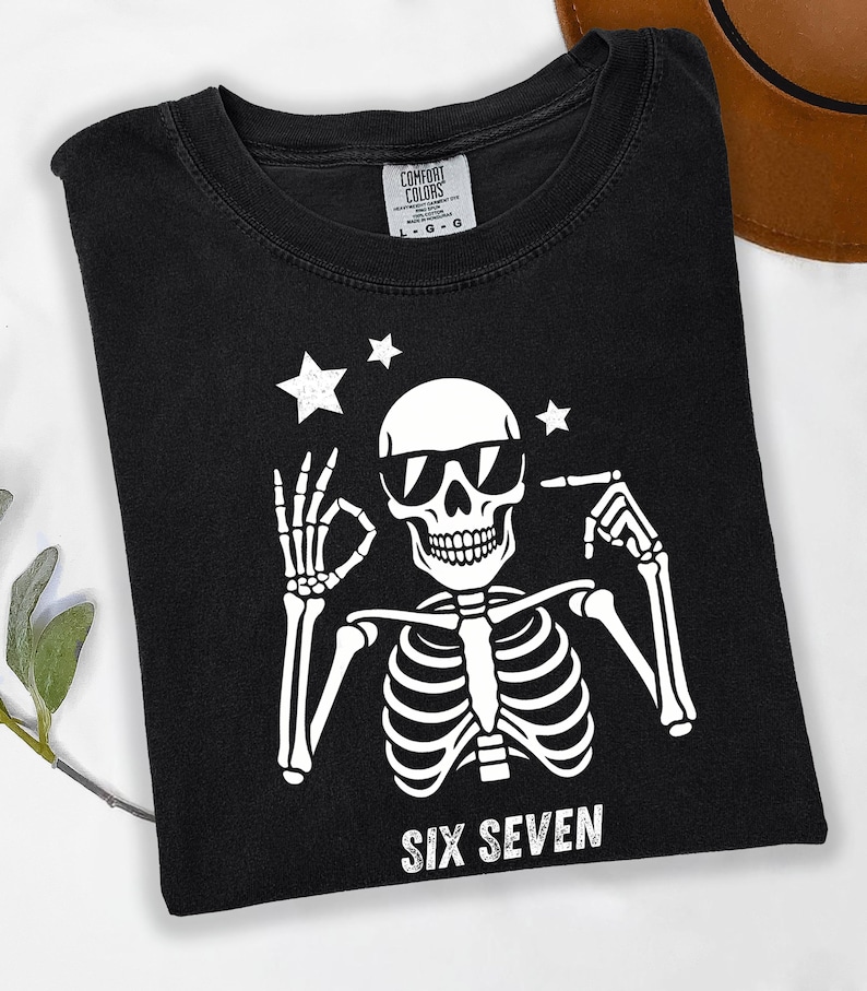 以下が含まれることがあります： サングラスをかけた白い骸骨のグラフィックが特徴の黒いTシャツ。「SIX SEVEN」の文字が骸骨の下にプリントされています。Tシャツはクルーネックで半袖です。