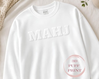 May include: A white crewneck sweatshirt featuring the text MAHJ in a raised 3D puff print across the chest. The garment has a Gildan brand label at the inner neckline and a circular graphic in the lower right corner with the text 3D PUFF PRINT.