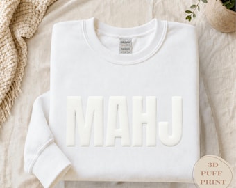 May include: A white crewneck sweatshirt featuring the letters MAHJ in a raised 3D puff print on the front. The garment has a ribbed collar, cuffs, and hem, with a Gildan 18000 brand label visible inside the neckline. A small circular tag reads 3D PUFF PRINT.