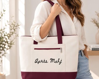 May include: A cream-colored canvas tote bag featuring a deep burgundy base and matching shoulder straps. The front includes a zippered pocket with the black text "Après Mahj" printed in a cursive script font centered on the pocket.
