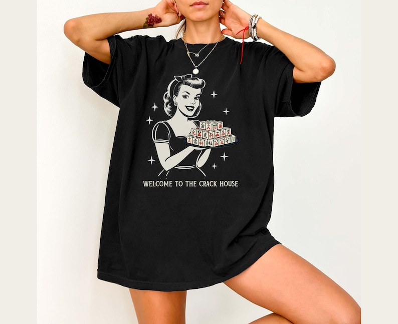 Op de afbeelding: Zwart oversized T-shirt met een retro-stijl afbeelding van een vrouw die een dienblad met items vasthoudt. De afbeelding bevat de tekst "WELCOME TO THE CRACK HOUSE" in het wit. Het shirt heeft korte mouwen en een relaxte pasvorm.