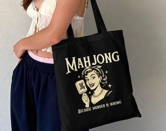 May include: A black tote bag featuring a cream-colored graphic of a person holding a mahjong tile. The text reads "MAHJONG" at the top and "BECAUSE MURDER IS WRONG" at the bottom, accented with small star shapes around the central figure.