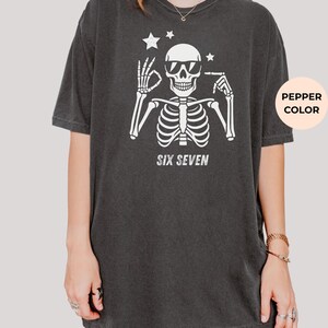以下が含まれることがあります： サングラスをかけた白い骸骨のグラフィックが特徴の濃いグレーのTシャツ。骸骨の下には「SIX SEVEN」の文字がプリントされています。骸骨の頭の上には2つの星があります。