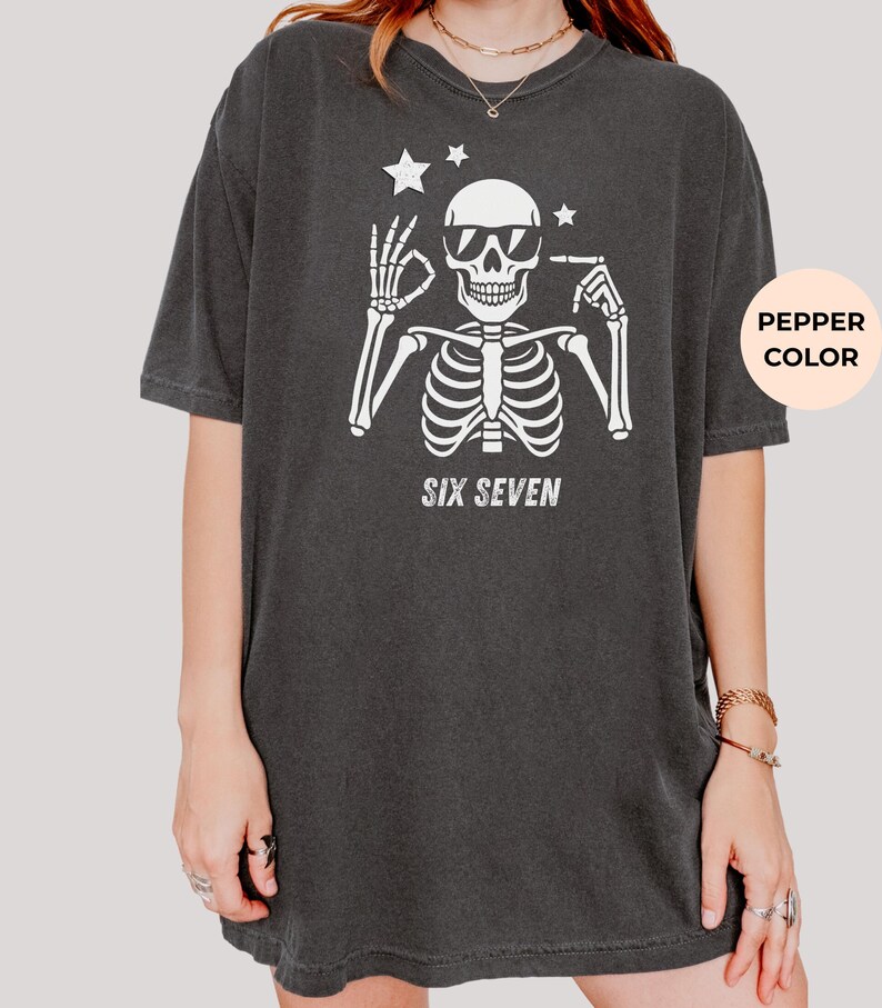 以下が含まれることがあります： サングラスをかけた白い骸骨のグラフィックが特徴の濃いグレーのTシャツ。骸骨の下には「SIX SEVEN」の文字がプリントされています。骸骨の頭の上には2つの星があります。