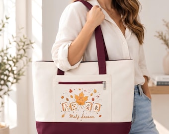May include: A cream-colored canvas tote bag with a deep burgundy base and matching shoulder straps. The front features a zippered pocket with a graphic of a smiling maple leaf and mahjong tiles, accompanied by the text Mahj Season.
