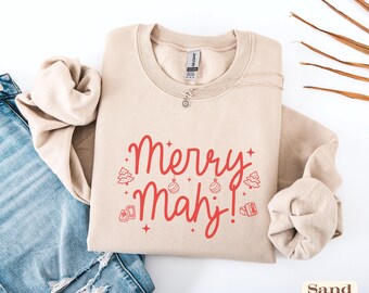 May include: A sand-colored crewneck sweatshirt featuring red text that reads Merry Mahj! The design includes small red illustrations of Christmas trees, ornaments, and mahjong tiles, accented with tiny stars. A silver chain necklace rests at the neckline.