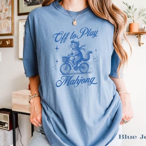 May include: A light blue t-shirt with a graphic of a cat riding a bicycle with a basket. The text reads "Off to Play Mahjong" in a script font. The shirt is a solid color.