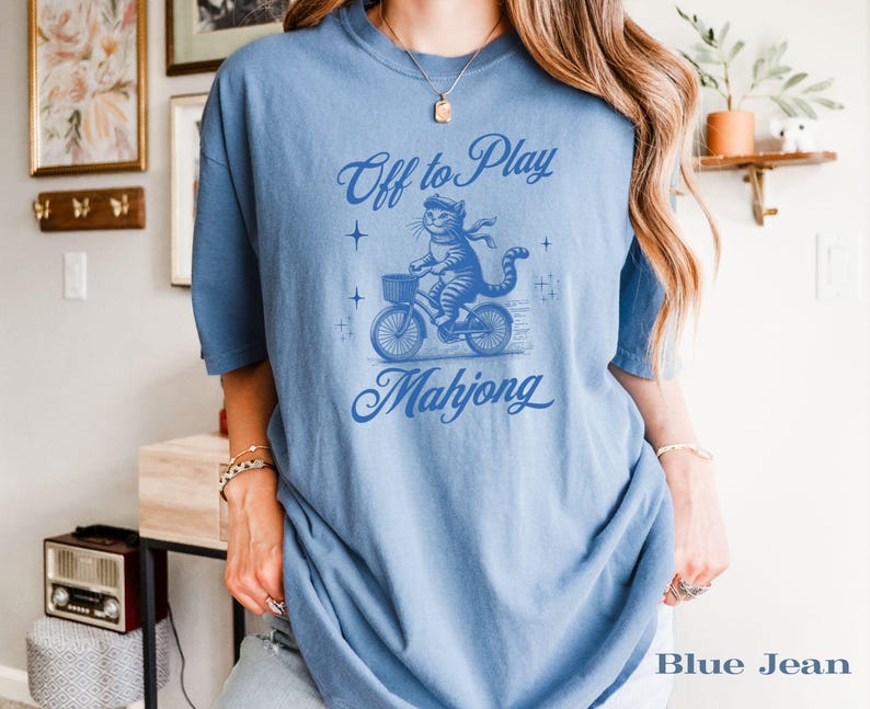 May include: A light blue t-shirt with a graphic of a cat riding a bicycle with a basket. The text reads "Off to Play Mahjong" in a script font. The shirt is a solid color.