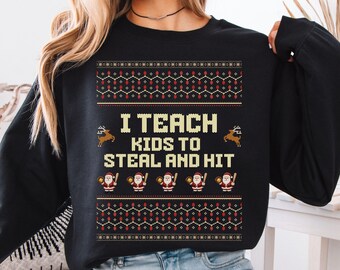 May include: A black long-sleeve sweatshirt featuring a holiday-themed graphic with the text "I TEACH KIDS TO STEAL AND HIT" in yellow, surrounded by illustrations of reindeer, Santa Claus figures holding baseball bats, and red light bulbs.