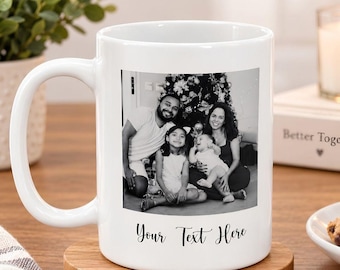 May include: A white ceramic mug featuring a black and white photograph of a family of four sitting in front of a Christmas tree. Below the photograph, the text "Your Text Here" is printed in a black cursive script font on the mug surface.
