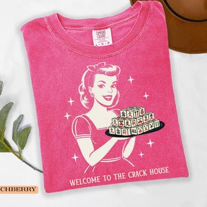Op de afbeelding: Een roze Comfort Colors t-shirt met een retro afbeelding van een vrouw die een dienblad met Mahjong-tegels vasthoudt. De tekst "WELCOME TO THE CRACK HOUSE" staat eronder. Het shirt heeft een ronde hals en korte mouwen.