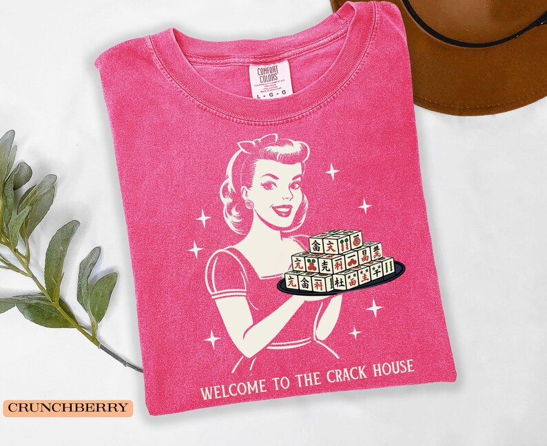 Op de afbeelding: Een roze Comfort Colors t-shirt met een retro afbeelding van een vrouw die een dienblad met Mahjong-tegels vasthoudt. De tekst "WELCOME TO THE CRACK HOUSE" staat eronder. Het shirt heeft een ronde hals en korte mouwen.
