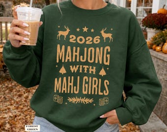 May include: A forest green crewneck sweatshirt featuring gold text that reads 2026 MAHJONG WITH MAHJ GIRLS. The design includes graphics of two deer, stars, two pine trees, a sprig of pine needles, and two mahjong tiles.