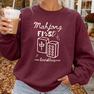 以下が含まれることがあります： マホガニー色のクルーネックトレーナー。クリーム色の文字で「Mahjong FIRST THEN Everything ELSE」とプリントされています。デザインには、漢字が描かれた麻雀牌とドットが描かれた麻雀牌の線画イラストが含まれています。