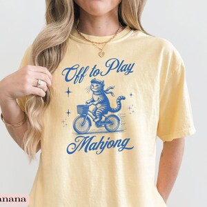 May include: Yellow t-shirt with a blue graphic of a cat riding a bicycle with a basket. The text "Off to Play Mahjong" is above and below the cat illustration. The shirt is a light yellow color.