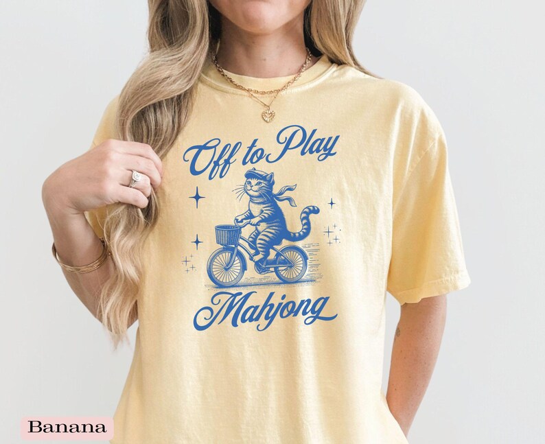 May include: Yellow t-shirt with a blue graphic of a cat riding a bicycle with a basket. The text "Off to Play Mahjong" is above and below the cat illustration. The shirt is a light yellow color.