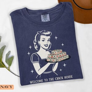 Op de afbeelding: Marineblauwe t-shirt met een retro-illustratie van een vrouw die Mahjong-tegels vasthoudt. De afbeelding bevat de tekst "WELCOME TO THE CRACK HOUSE" en sterren. Het shirt is gemaakt van zacht materiaal.