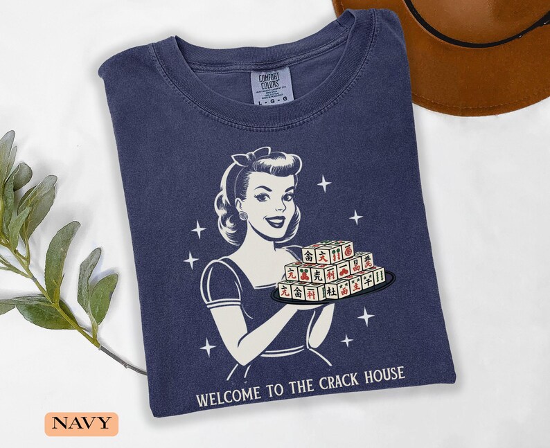 Op de afbeelding: Marineblauwe t-shirt met een retro-illustratie van een vrouw die Mahjong-tegels vasthoudt. De afbeelding bevat de tekst "WELCOME TO THE CRACK HOUSE" en sterren. Het shirt is gemaakt van zacht materiaal.