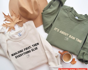 May include: A cream sweatshirt with black text reading "MAHJONG FIRST, THEN EVERYTHING ELSE" and an olive green sweatshirt with beige text reading "IT'S ABOUT BAM TIME," both featuring small mahjong tile graphics, a tan hat, and a mug.